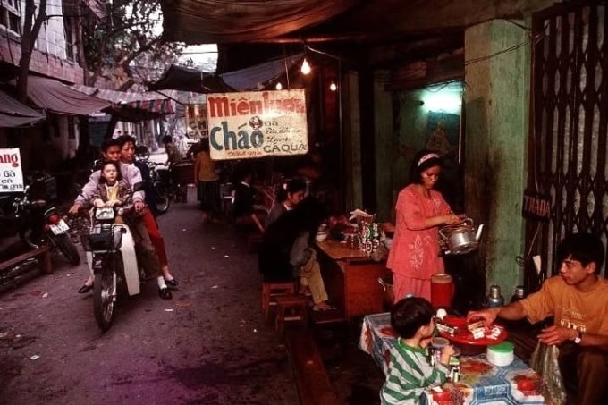 Người dân lái xe máy Honda len vào một quán ăn nhỏ trong ngõ. Quán bày bàn ghế đơn giản, thắp sáng bằng vài bóng đèn tròn, phía trên có tấm bạt nilon và biển hiệu viết tay. Hai bên là các ngôi nhà tập thể cũ, hình ảnh quen thuộc của phố phường Hà Nội thời trước.