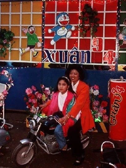 Người dân tạo dáng chụp ảnh Tết trên xe máy, trước không gian trang trí rực rỡ hoa giả. Phía sau là hình hai nhân vật Doraemon và Nobita trong bộ truyện Doraemon của Nhật Bản. Tác phẩm lần đầu xuất bản ở Việt Nam năm 1992, gắn bó với thiếu nhi nhiều thế hệ.